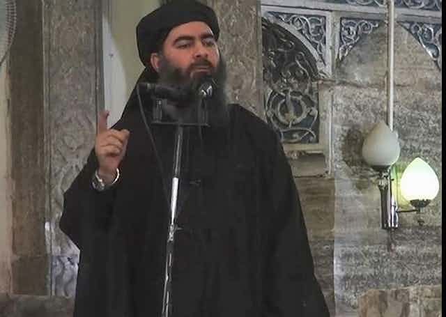 Bearded man dressed in black and wearing a turban speaks into a microphone.