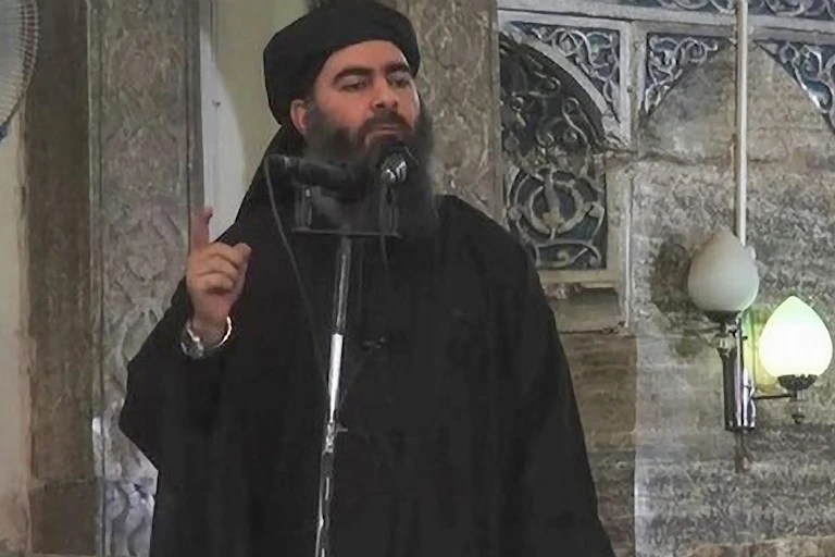 Bearded man dressed in black and wearing a turban speaks into a microphone.