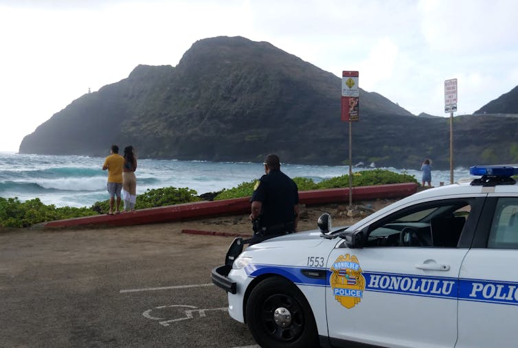 Police car from Honolulu police parked in a parking lot near the beach with an officer standing in front of it; in the backgroundn a couple looks out at the ocean