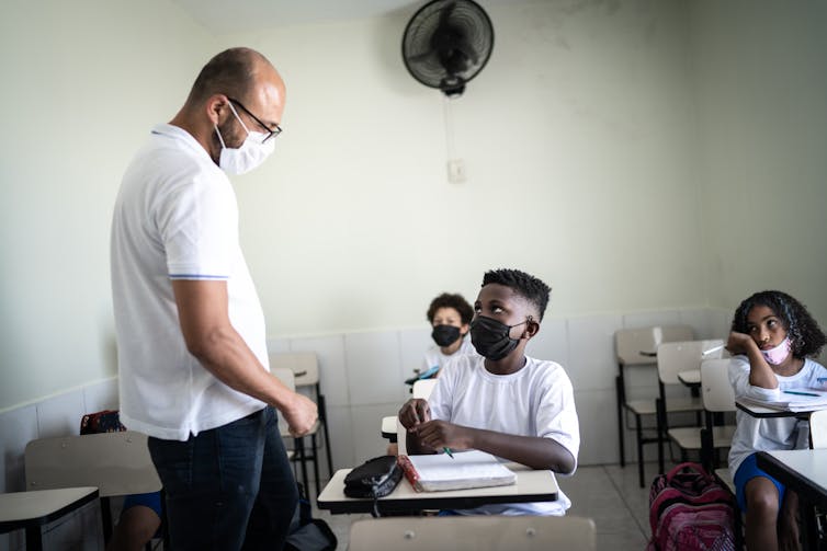 A teacher wearing a mask walks up to a student in class working on an assignment.