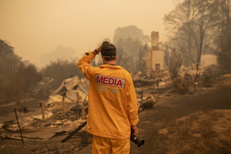 A man in a yellow shirt reading MEDIA looks at a property destroyed by fire.