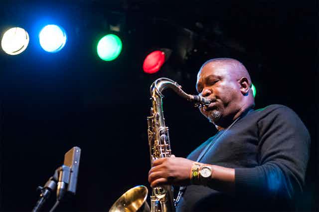 Under a row of multi-coloured stage lights, a man blows into a saxophone. In a plain black jersey, his eyes are closed as he plays.
