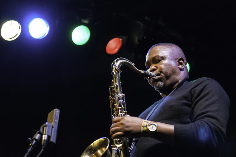 Under a row of multi-coloured stage lights, a man blows into a saxophone. In a plain black jersey, his eyes are closed as he plays.