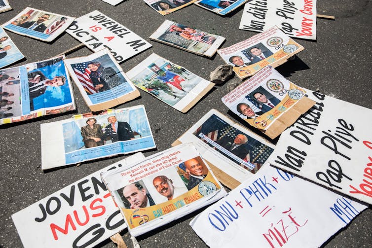 Placas de protesto vistas caídas no chão, dizendo "Jovenel deve ir" em inglês e crioulo