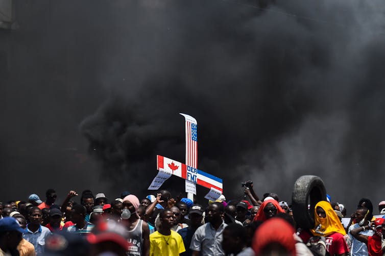 Crowd in the street under smoky skies hold up a sign with U.S., Canadian and other foreign flags