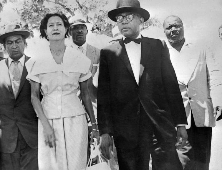 Black-and-white image of François Duvalier, in a suit, and his wife, in a dress, surrounded by watchful men