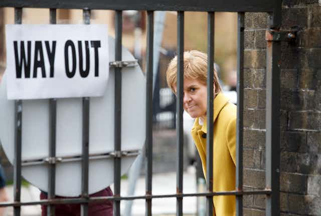 Nicola Sturgeon looks out from behind a gate with a 'way out' sign on it.