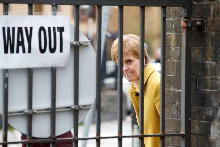 Nicola Sturgeon looks out from behind a gate with a 'way out' sign on it.