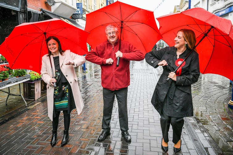 Mark Drakeford smiles an bumps elbows with two women, one with a red rosette, all carrying red umbrellas.