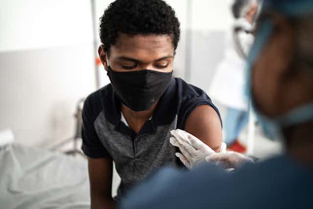 A teenage boy wearing a mask watches a nurse prepare his arm for the vaccine