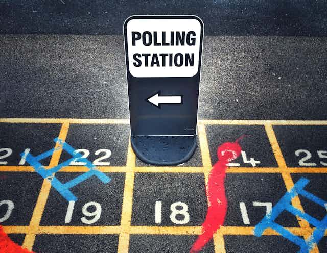 A sign for a polling station sitting on top of a snakes and ladders game grid painted on a playground.