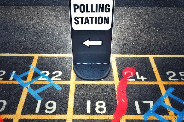 A sign for a polling station sitting on top of a snakes and ladders game grid painted on a playground.