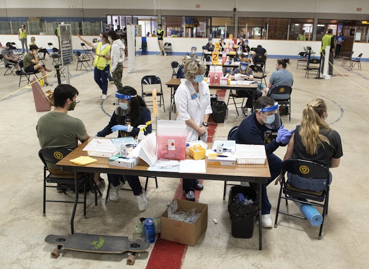 Students and medical personal at tables in a large open area.