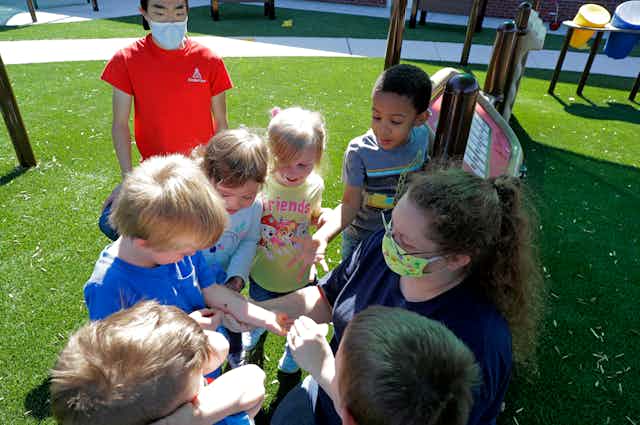 daycare teachers wearing masks work with kids examining a spider they found on a playground in the background