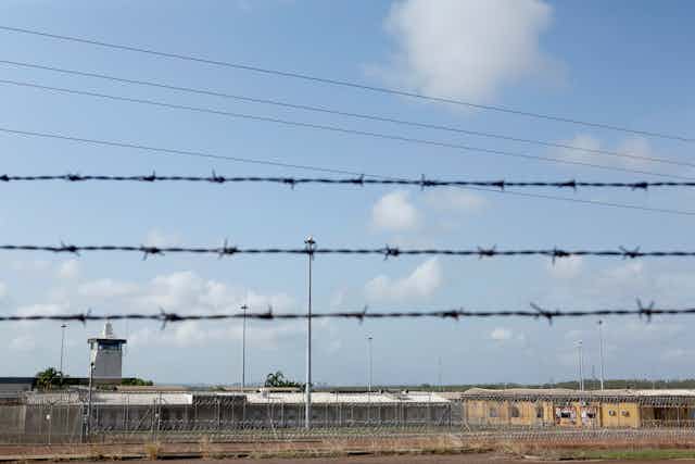 A barbed wire fence in focus, with the Don Dale Youth Detention Centre in the background.