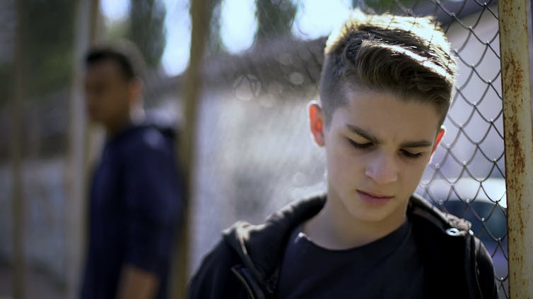 A teenage boy leans against a fence, appearing despondent.