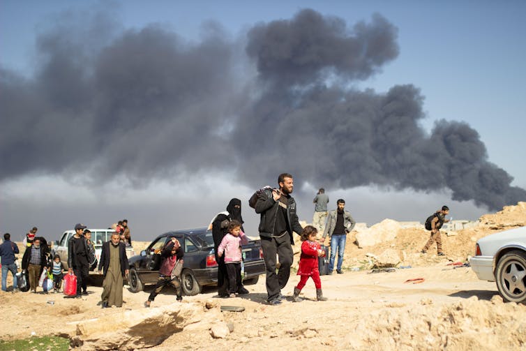 People walking carrying belongings with black smoke behind them.