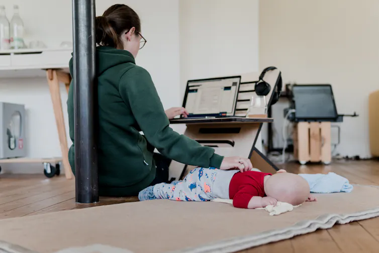 A woman sits on the ground with one hand on a sleeping baby and the other typing at a laptop