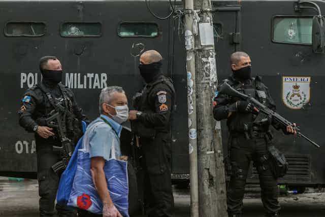 A man in a COVID mask walks past masked Brazilian security forces in the Jacarezinho favela in Rio, May 2021.
