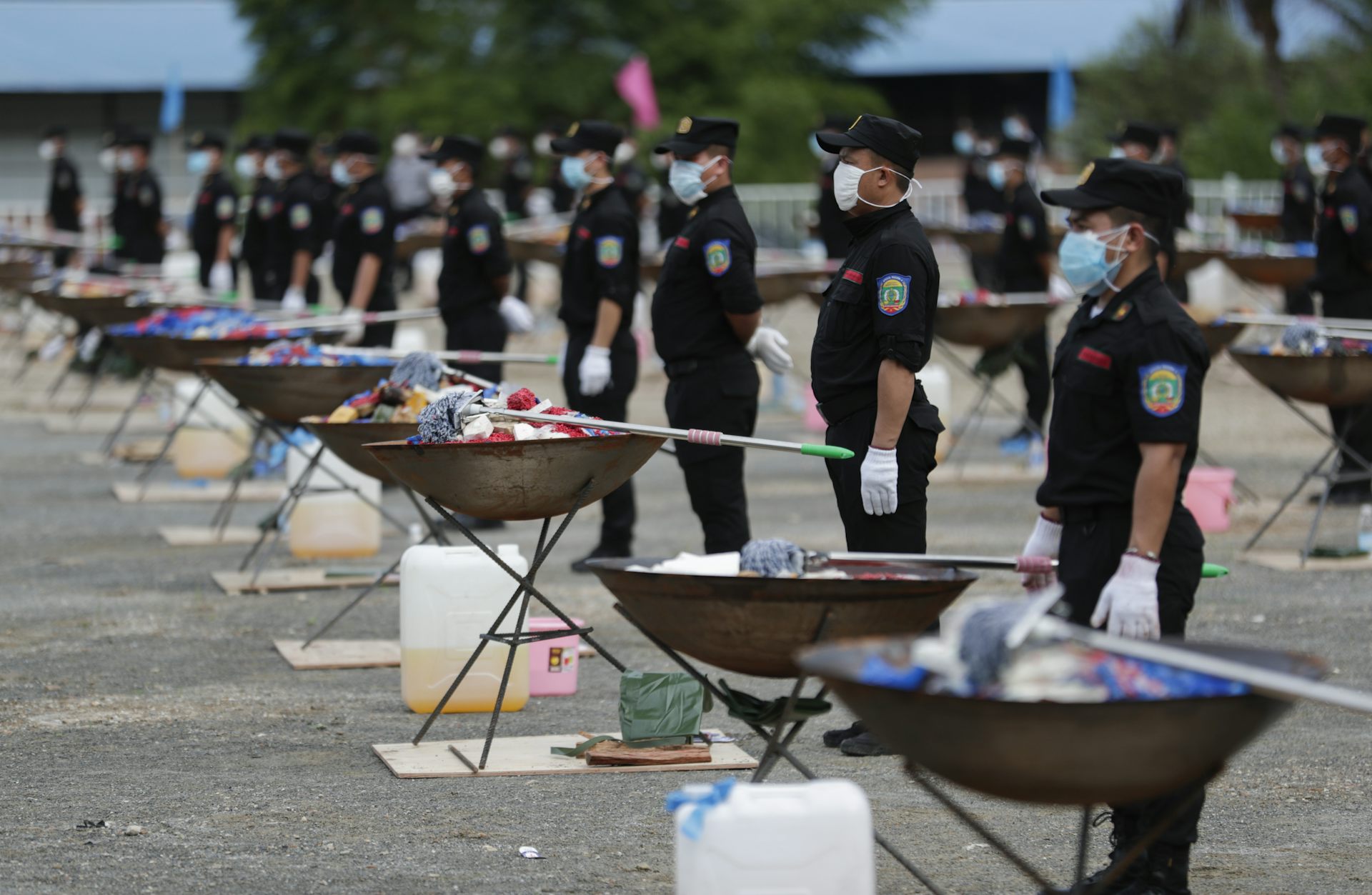 Masked police officers standing with braziers filled with narcotics, preparing to burn the drugs. On the ground by the braziers are jerrycans with petrol.