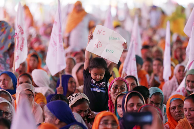 A young boy in a black t-shirt surroundd by women in traditional dress waves a placard reading 'No farmers, no food' at a rally in India, April 2021.