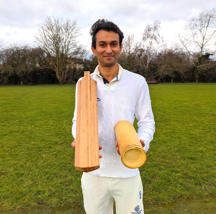 A man in cricketing clothes holds a bat and a piece of bamboo