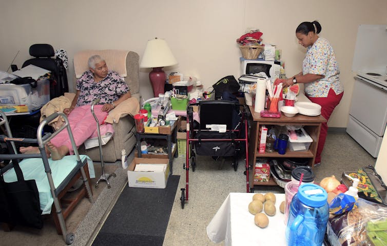 An elderly woman lies in a bed as a home care aide prepares some food.