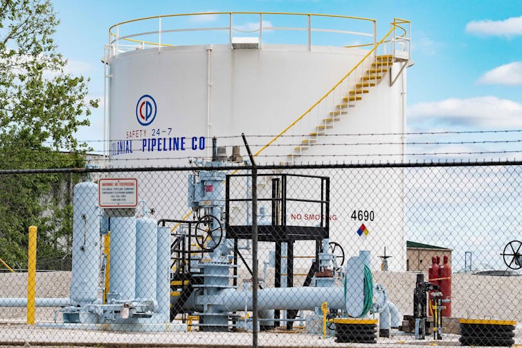 A chain-link fence topped with barbed wire in the foreground, large pipes and valves in front of a large white storage tank labeled Colonial Pipeline Co