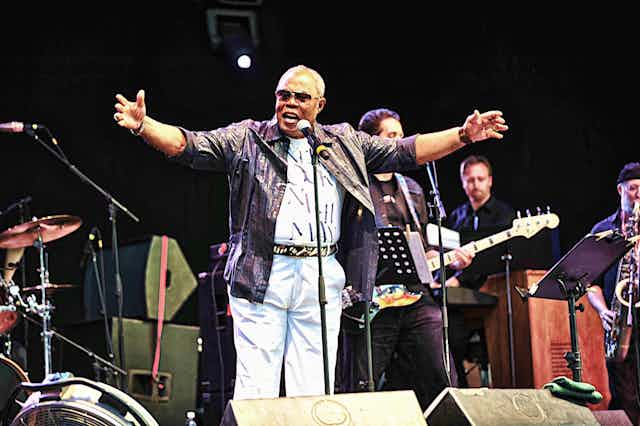 Musician Sam Moore stands behind a microphone wearing sunglasses with his arms raised