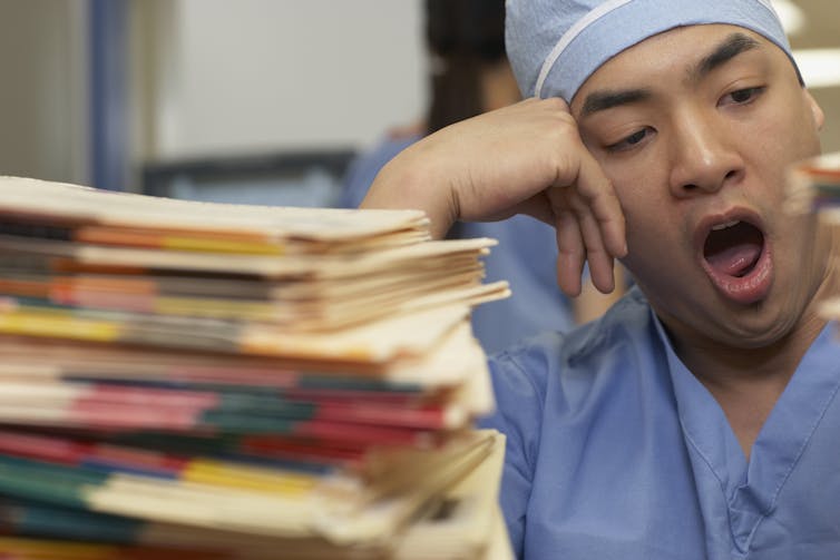 health care worker yawning next to stack of medical records