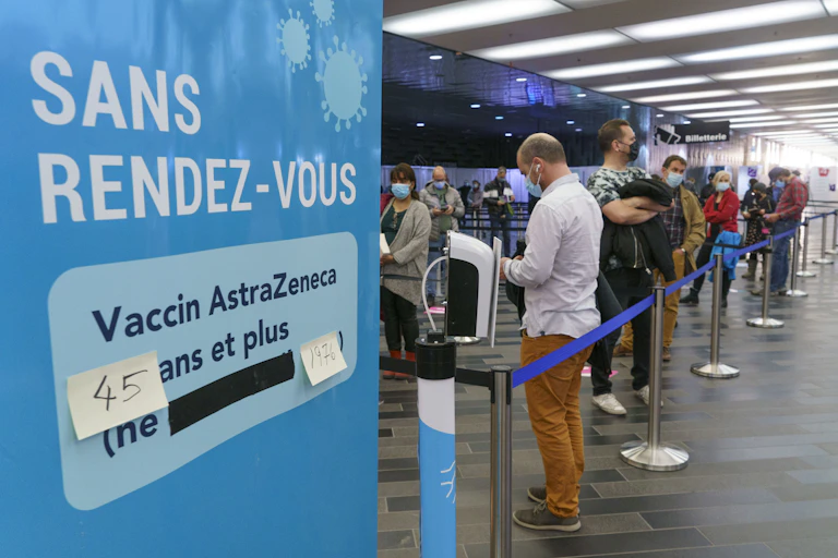 Des gens portant un masque font la file pour être vaccinés. À L'avant-plan une affiche bleue indique que le vaccin d'AstraZeneca est disponible pour les 45 an et plus.