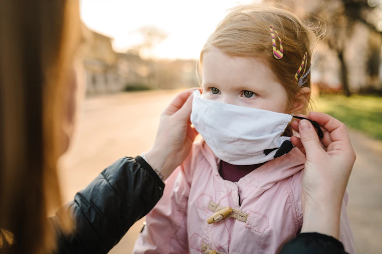 Una mujer pone una mascarilla a una niña pelirroja.