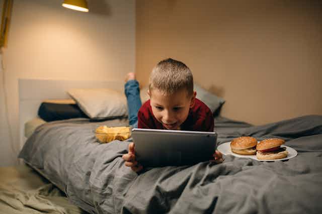 Un niño sobre una cama con una tablet, dos hamburguesas y cuenco de patatas fritas.