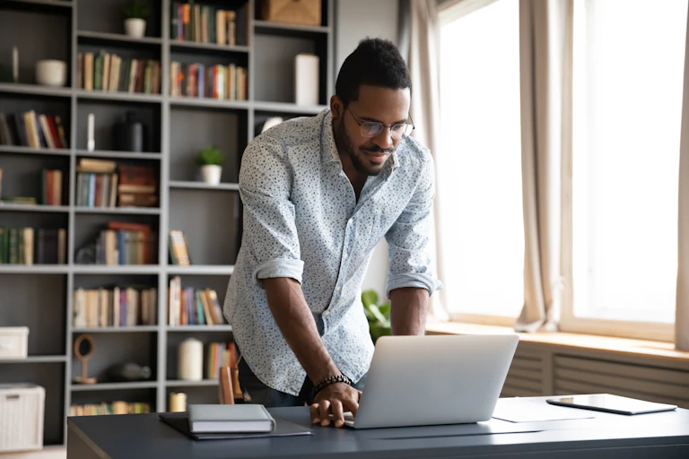 Young man stands at his desk at home, working on his laptop.