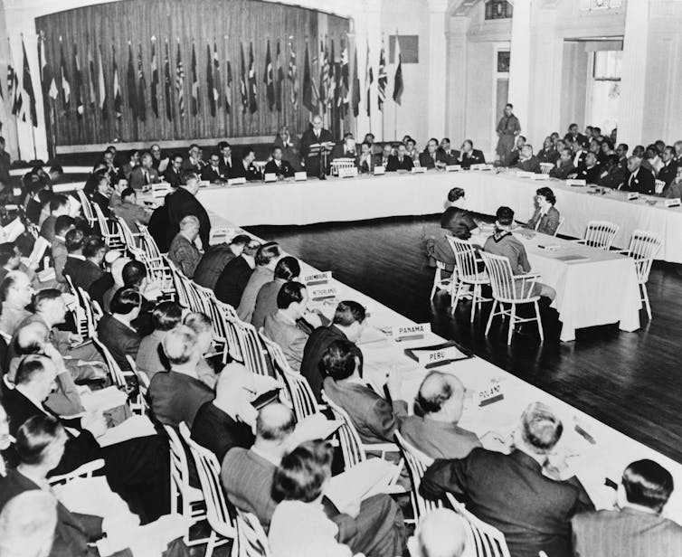 Black and white photo of delegates seated around horseshoe table with flags in the background.