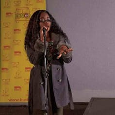 Against a yellow backdrop, a woman with long hair and wearing agree coat holds a microphone, her other hand held up with fingers splayed.