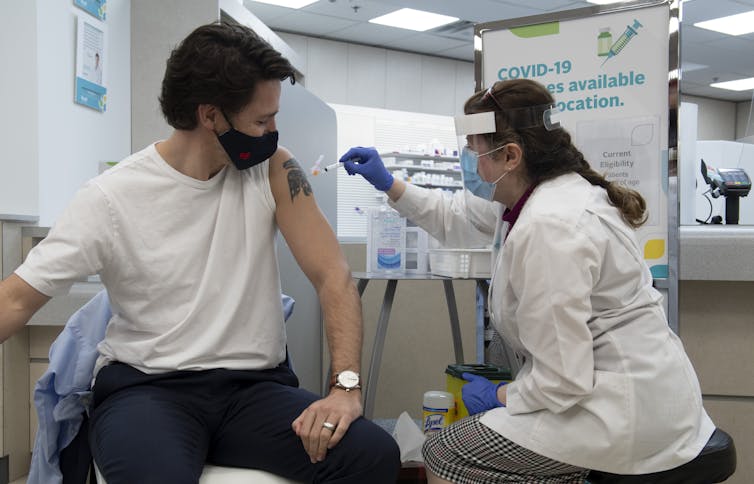Le premier ministre du Canada, Justin Trudeau, la manche de son t-shirt blanc relevée, est vacciné par une femme en sarrau blanc
