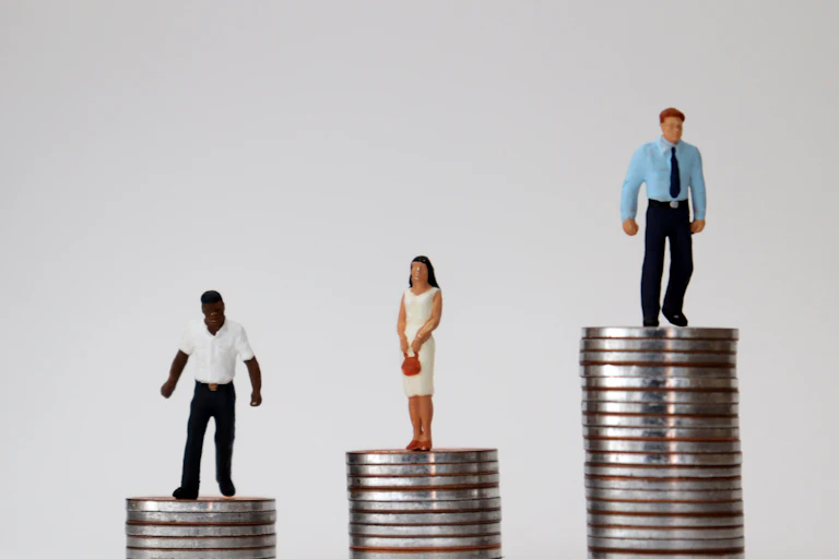 Miniature figures of different races and genders standing on piles of coins of varying heights