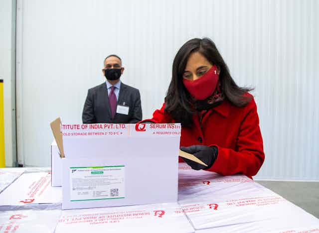 Minister of Public Services Anita Anand wearing a red face mask, opening a box of vaccine.