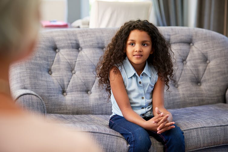A young girl sits on a couch, appears to be talking with a therapist.