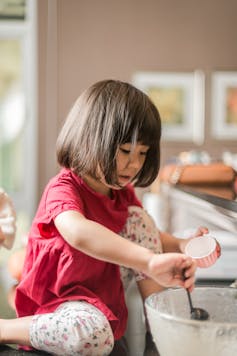 A little girl in a red dress sits on a counter top stirring cake mixture to make cupcakes