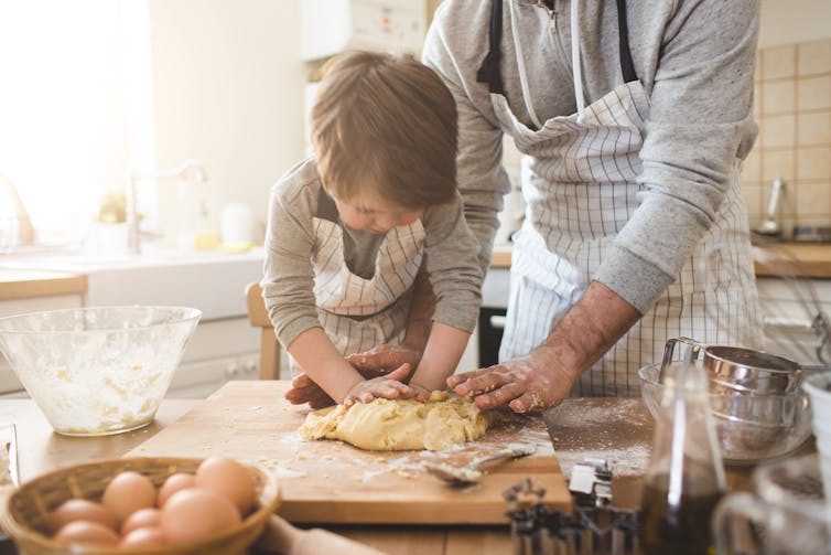A father teaches his son how to kneed dough on a kitchen surface