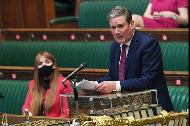 Keir Starmer speaking in the House of Commons while Angela Rayner looks on from the background.