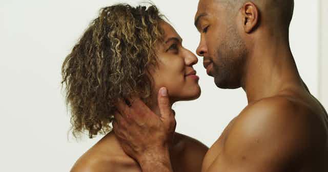 A man and a woman, their bare shoulders revealed, face one another, their lips close as if they are about to kiss.