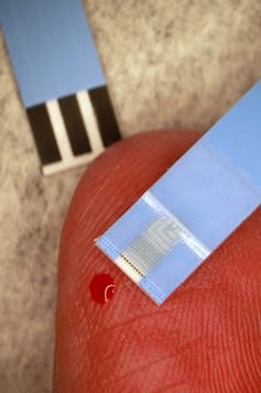 A person's finger with a small drop of blood and a strip of blue paper.