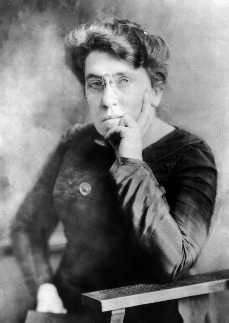 Emma Goldman in 1911 with dark hair in a bun, seated, looking straight at the camera through rimless spectacles.