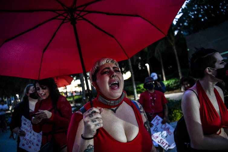 People, including one under a red umbrella, march and shout in the street