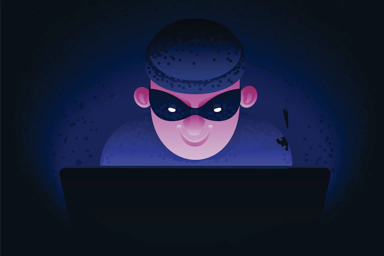 cartoon image of a man wearing a dark knit cap and a mask over his eyes grinning as he uses a laptop