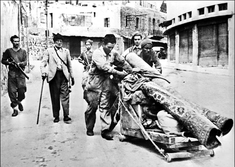 A black-and-white image shows two men pushing a wooden cart of belongings through a city street. An armed man follows them.