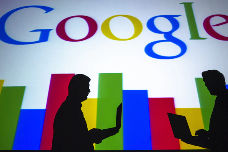The silhouettes of two people using laptops are seen against the Google logo.